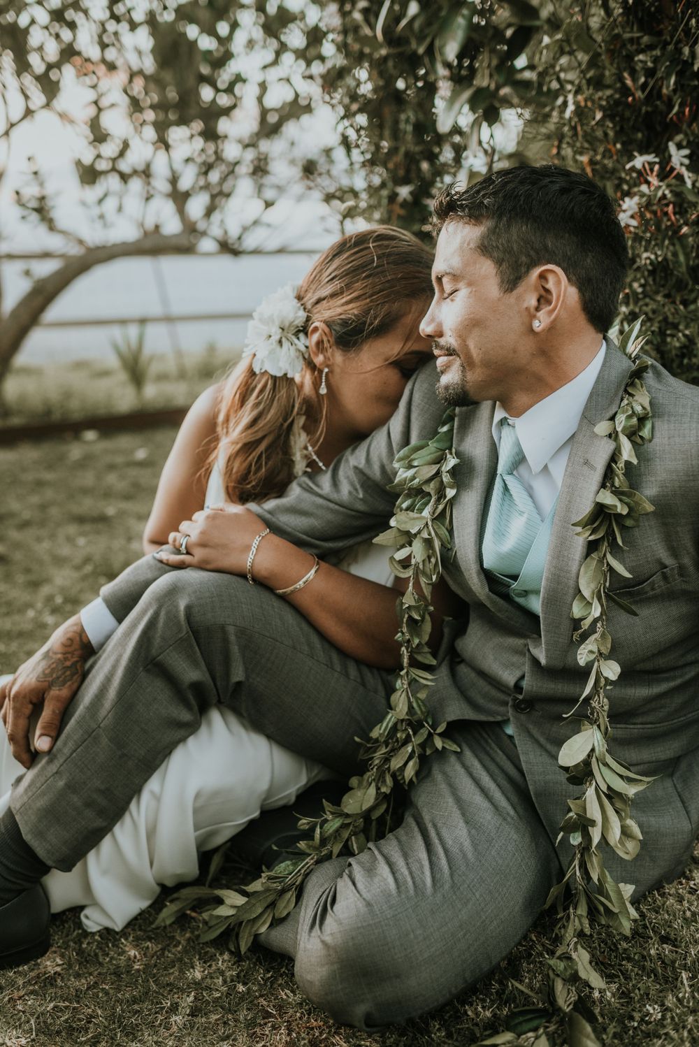 Couple wearing green leaf leis embrace while sitting together outdoors.