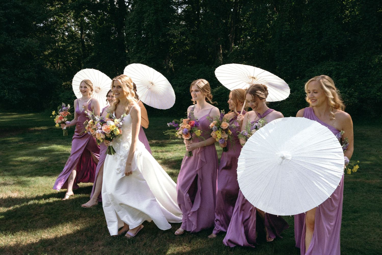 A wedding party in purple dresses holds white parasols while walking through a sunlit garden setting.