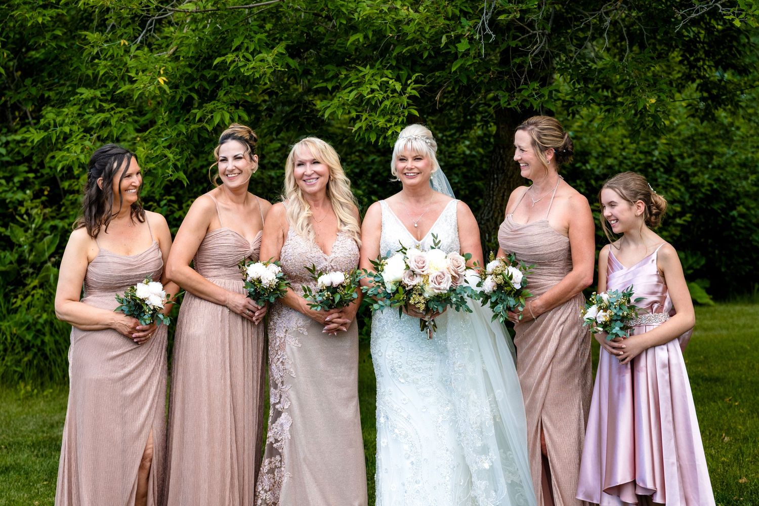 A group of bridesmaids in dusty rose dresses pose together with the bride in white at an outdoor garden wedding.