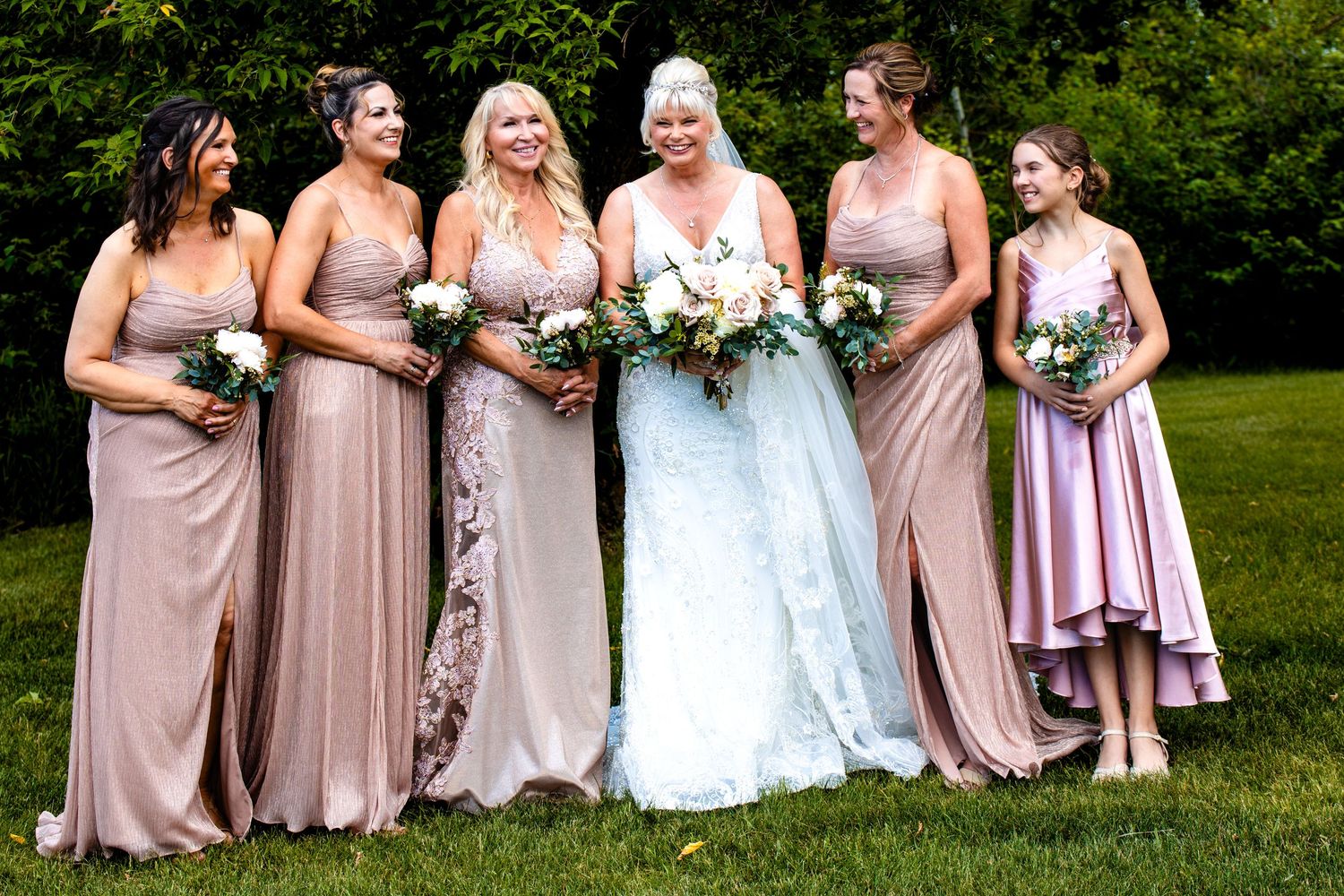 Wedding party in blush pink dresses poses together on grass holding floral bouquets.