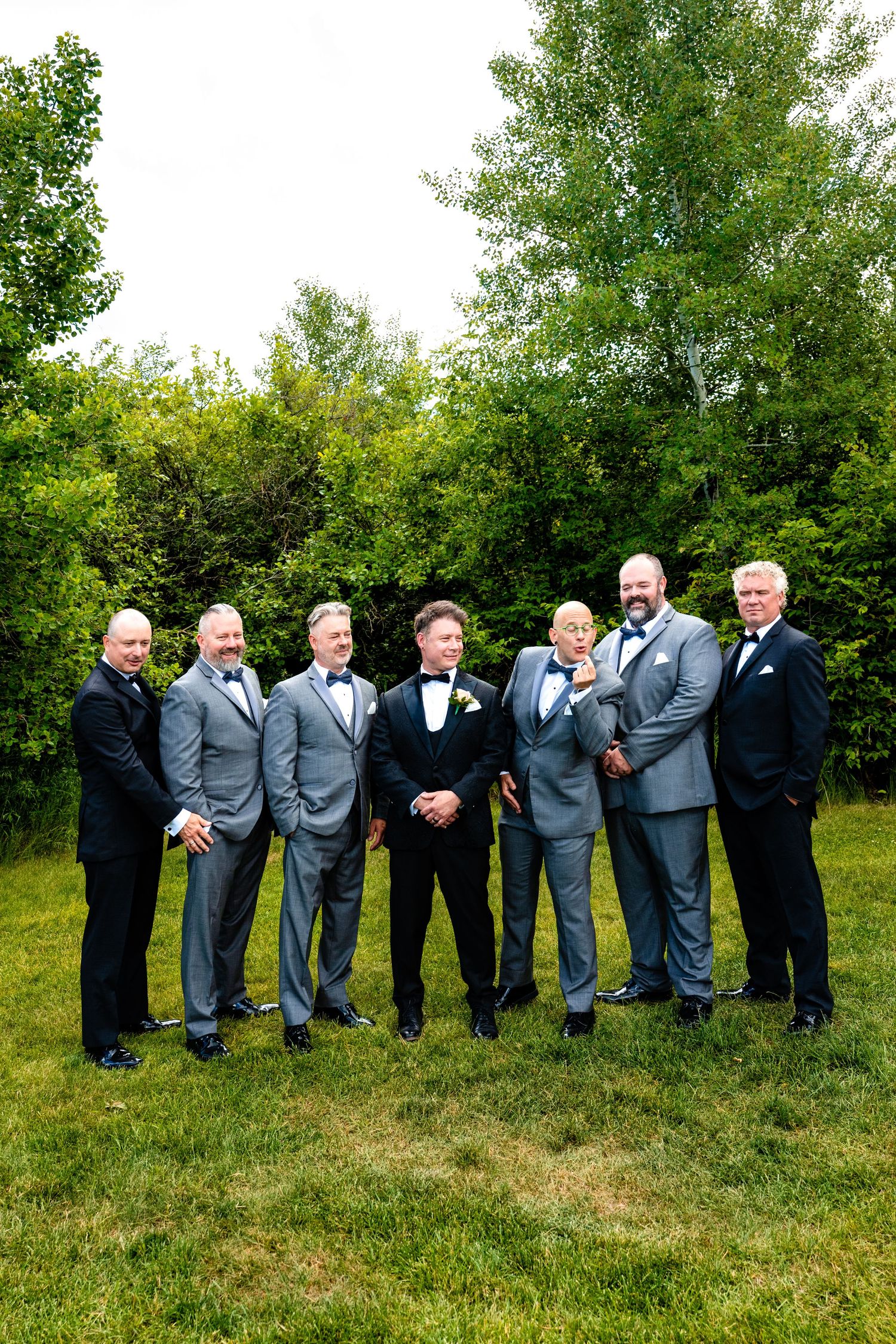Groomsmen in matching gray suits and black bowties stand in a line on green grass with trees in background.