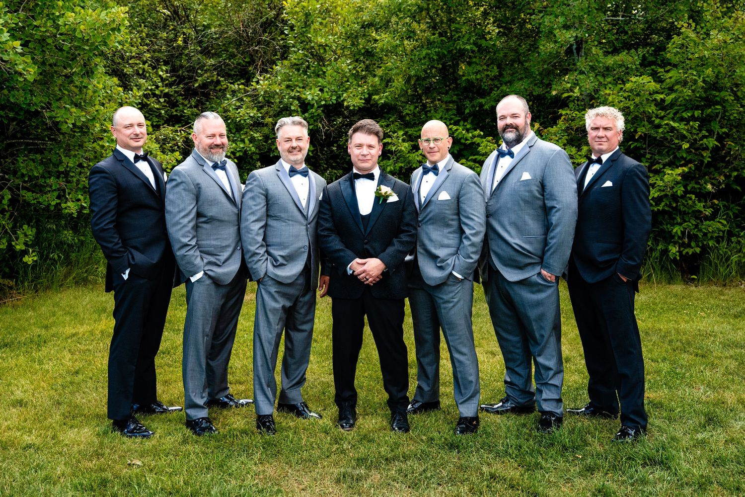 Wedding party in gray suits poses together outdoors in front of green foliage at a summer celebration.