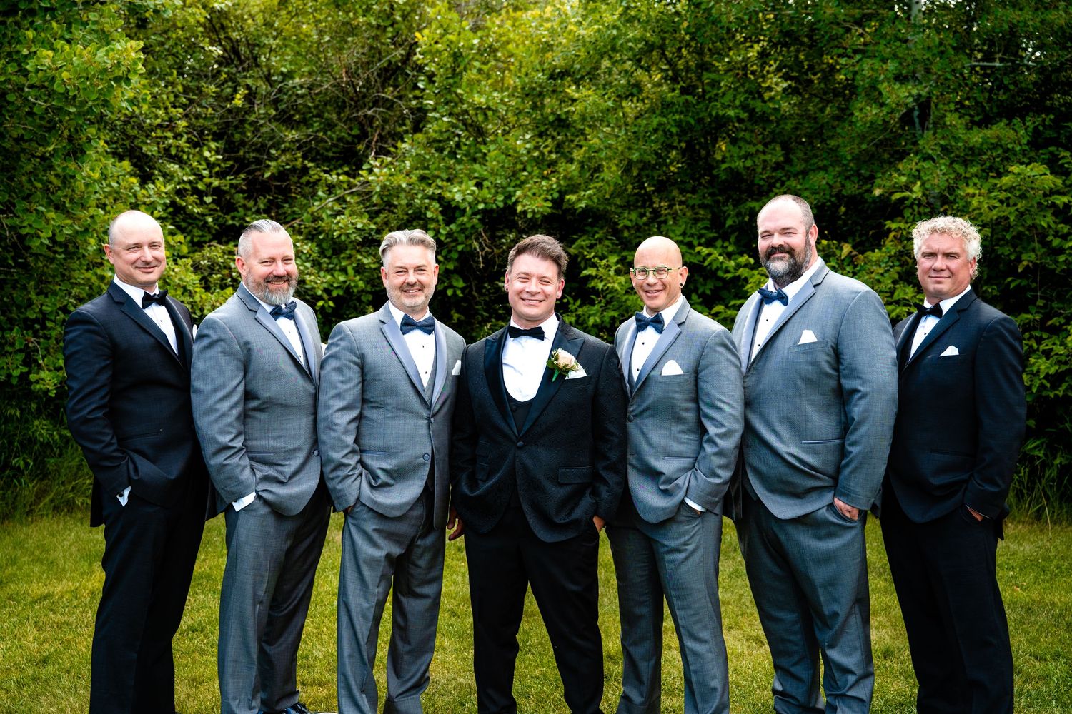 A group of groomsmen in gray and black suits pose together on a grassy lawn during a wedding celebration.