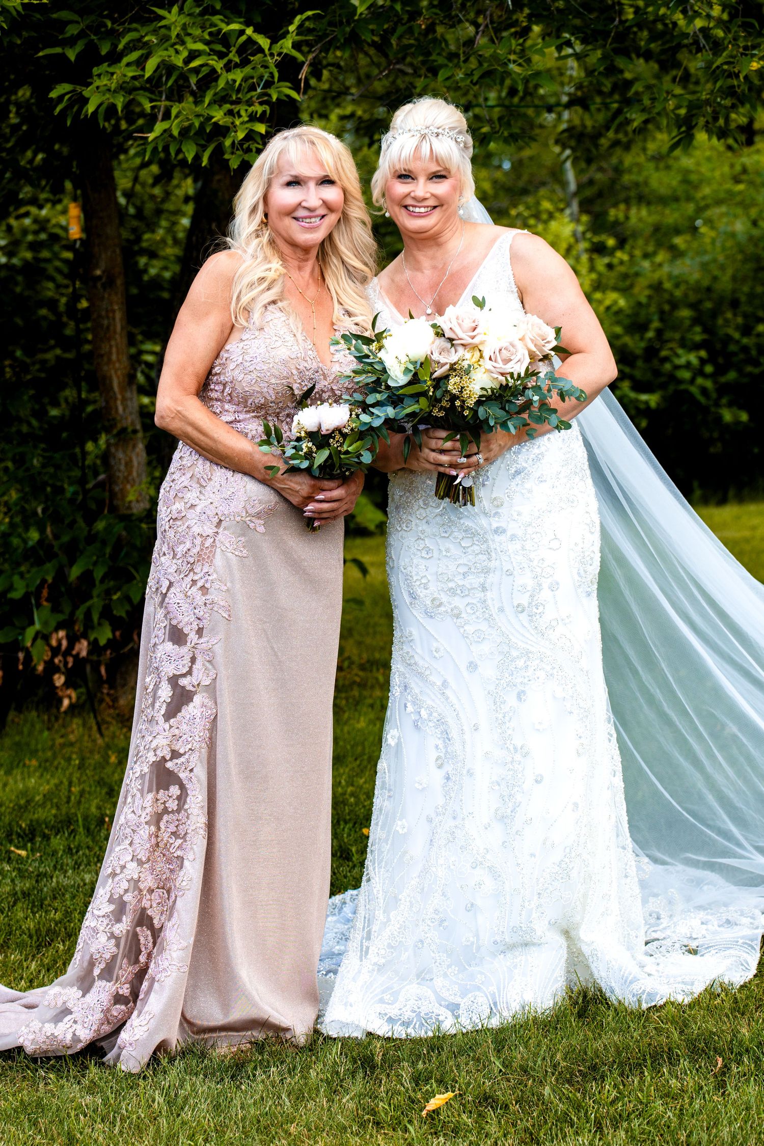 Two people in elegant dresses pose together on a grassy lawn during a wedding celebration.