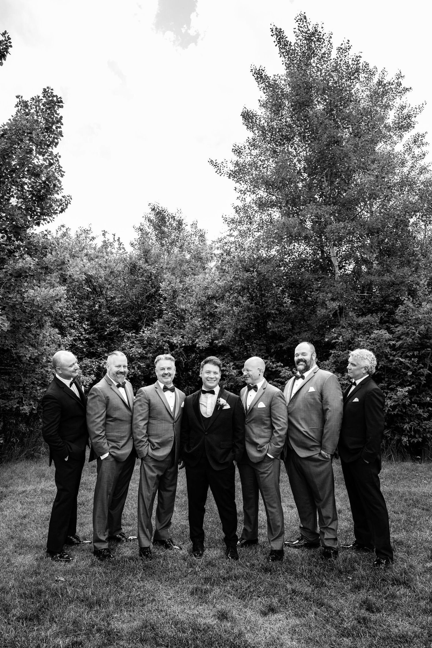 A black and white photo showing groomsmen lined up outdoors in formal attire against a natural backdrop.