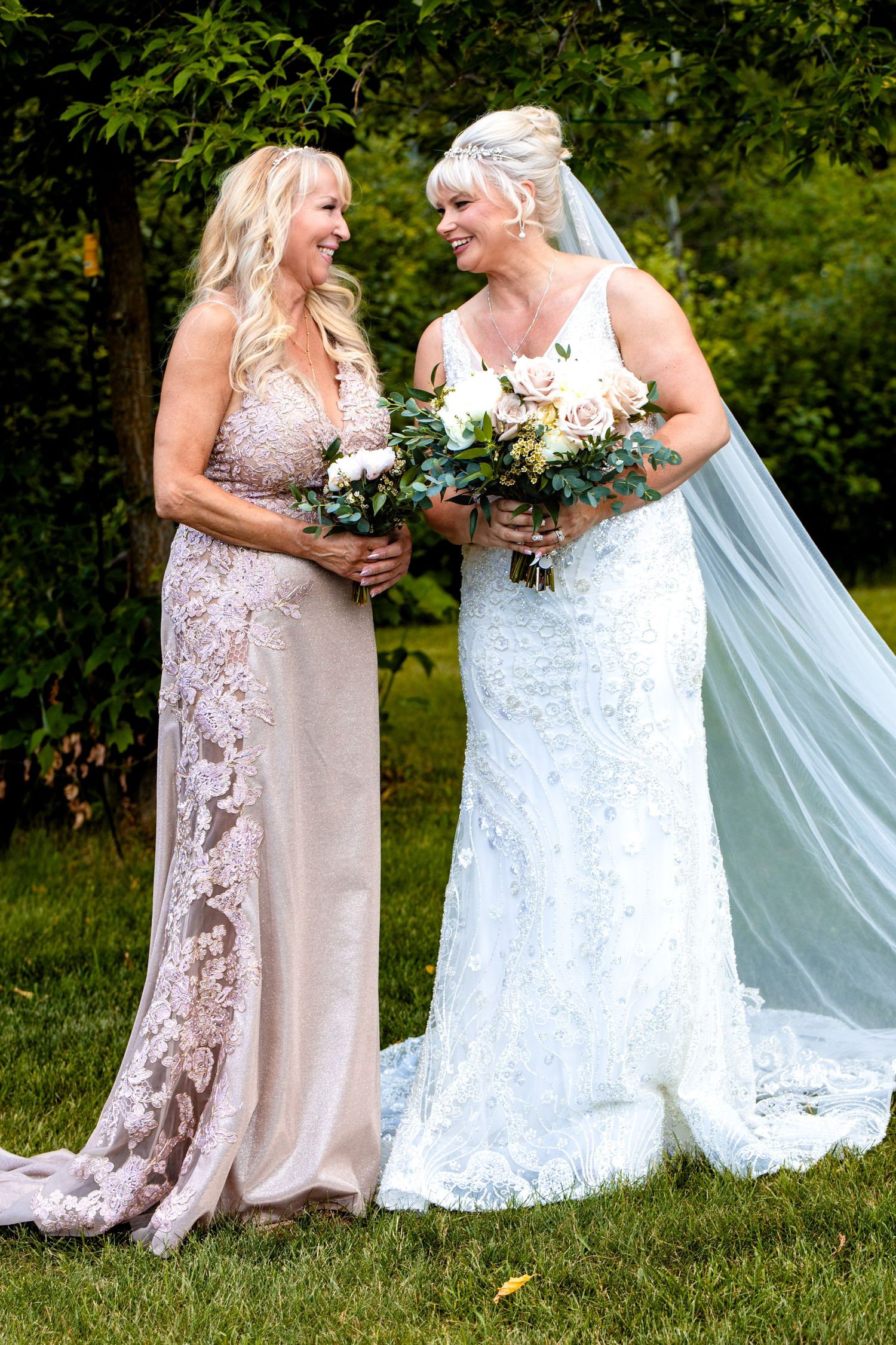 Two people in formal dresses share a joyful moment on grassy lawn with one wearing white lace and holding bouquet.