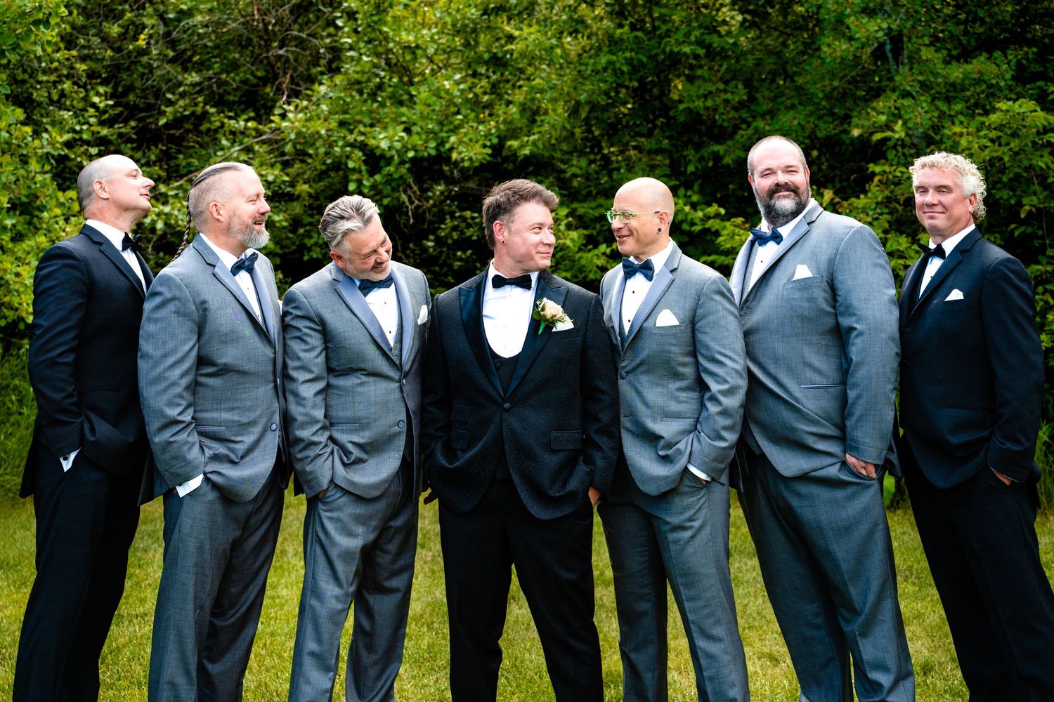 Groomsmen in gray suits share a candid laughing moment together in a line against green foliage background.