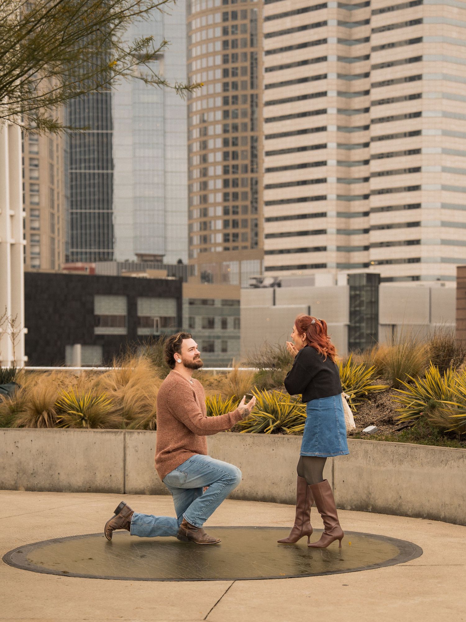 A couple spins playfully in a circular concrete area with city buildings in the background.