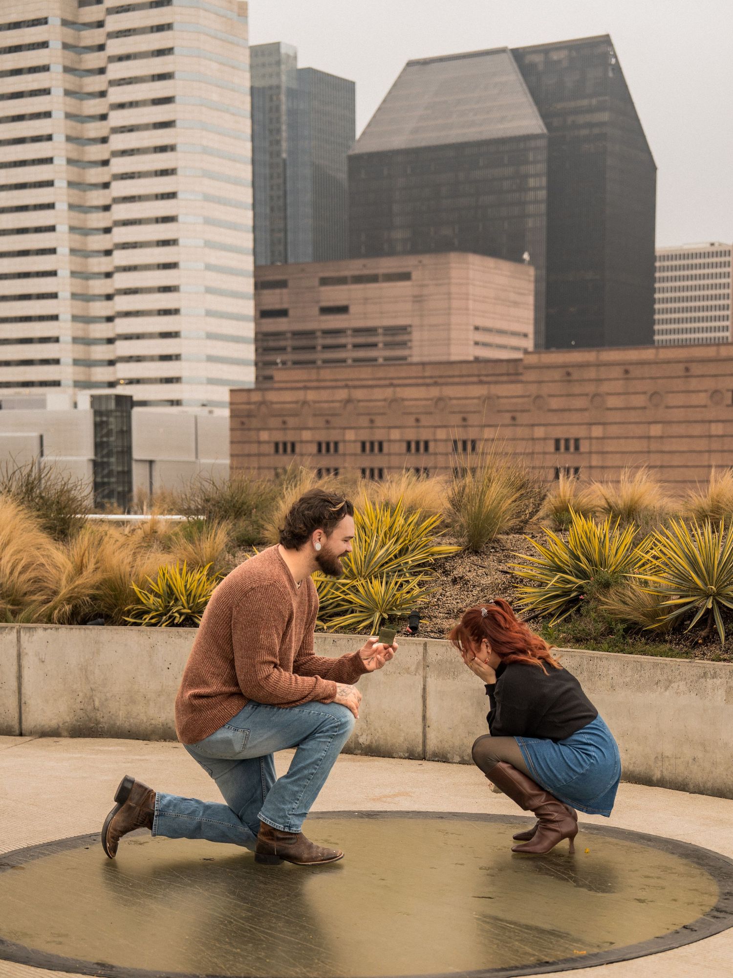 A couple sharing a romantic proposal moment on a rooftop garden with Houston's downtown skyline in the background.