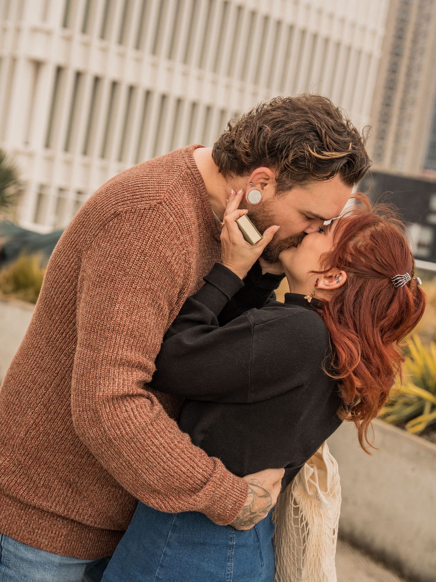 A romantic couple embraces against a backdrop of urban architecture and railings.