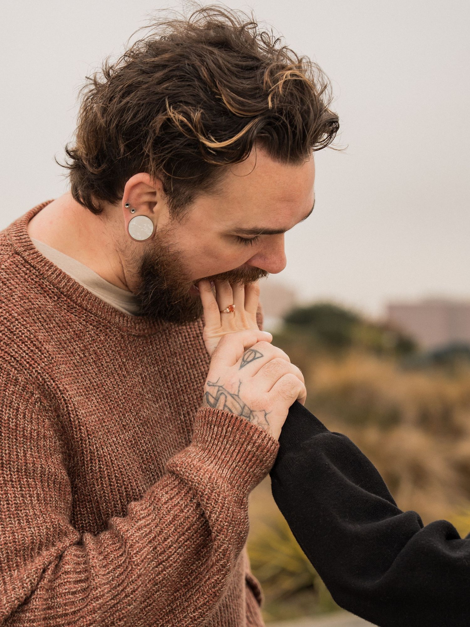 Person in brown sweater kissing someone's hand with emotion against an urban backdrop.