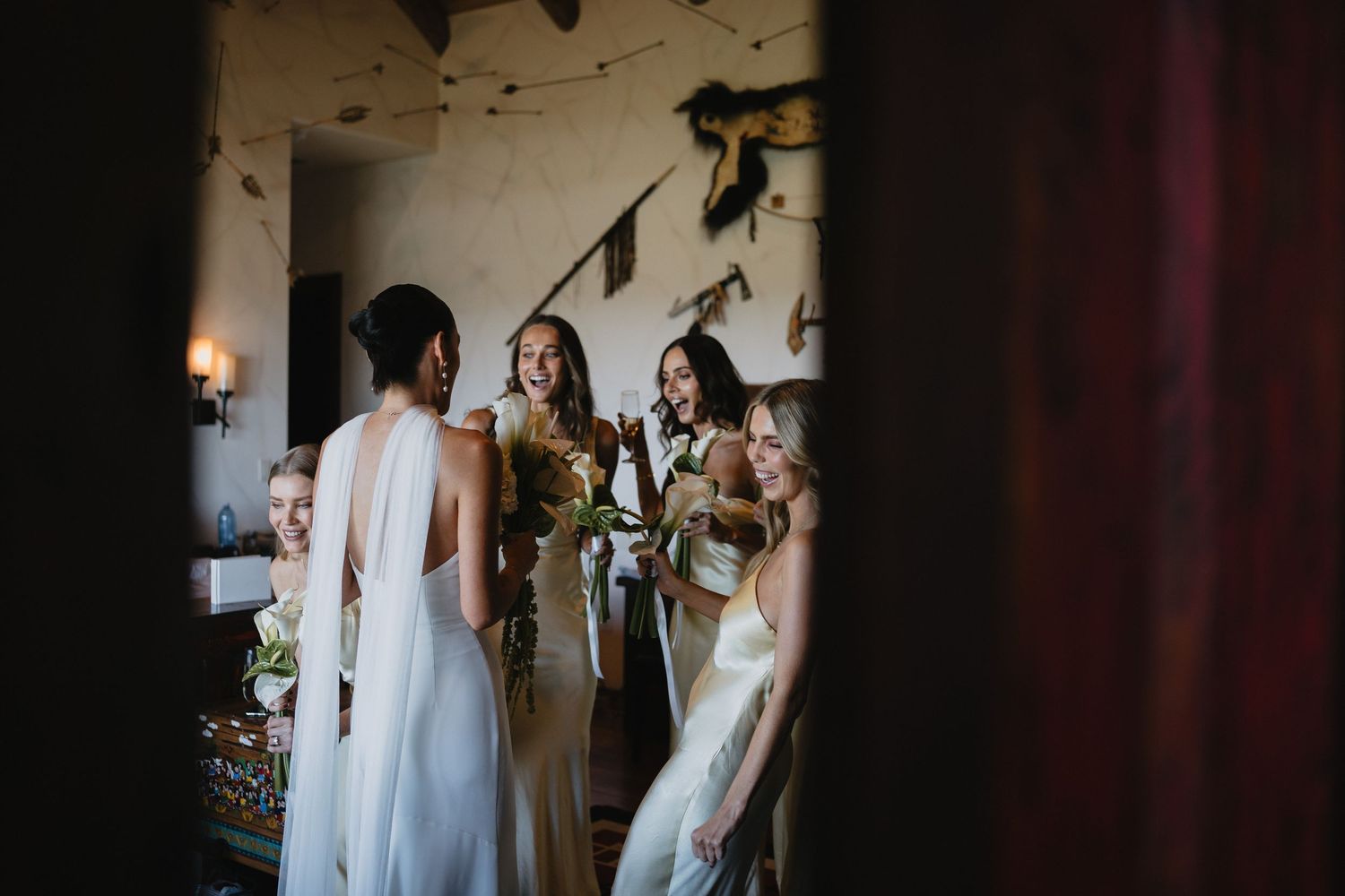 Bride shares a joyful moment with smiling bridesmaids holding bouquets inside a warmly lit rustic venue.