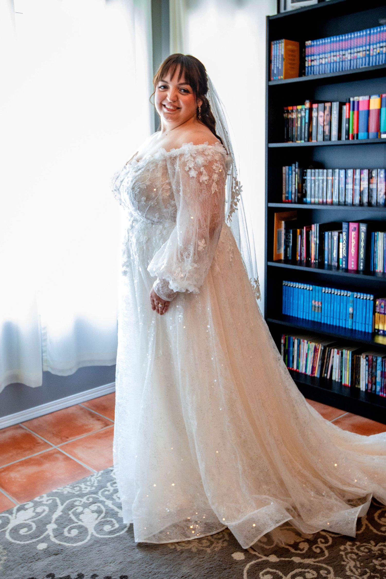 A bride poses in a stunning long-sleeve lace wedding gown with sparkle details against a bookshelf backdrop.