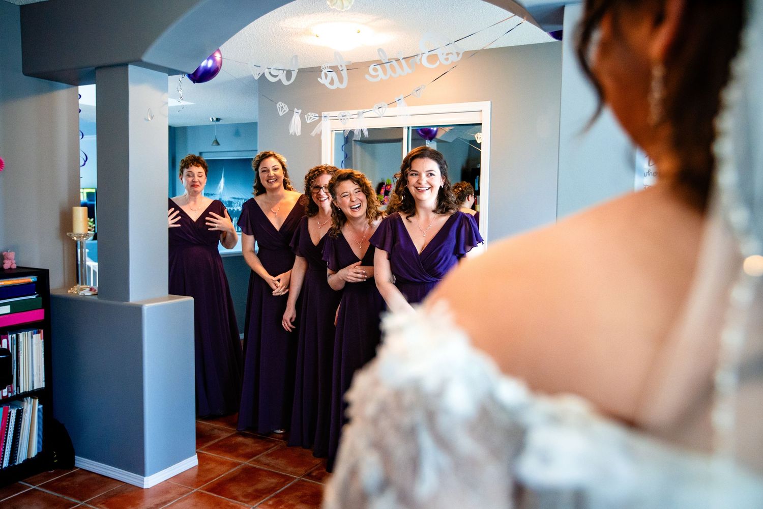 A bridal party gets ready in a blue-walled room with bridesmaids in purple dresses helping with preparations.