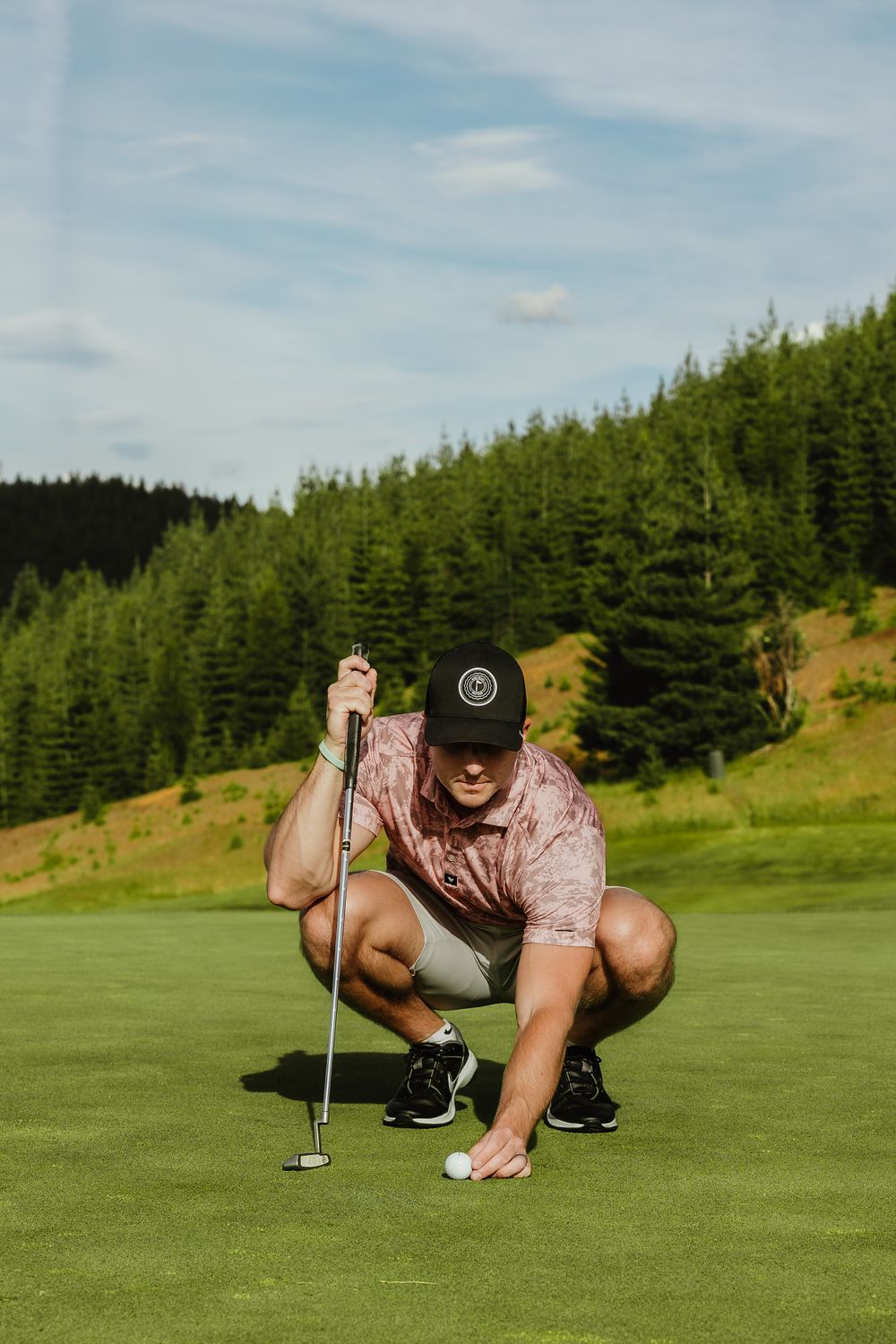 A golfer crouches on a green course analyzing the putting line while surrounded by evergreen trees on a sunny day.