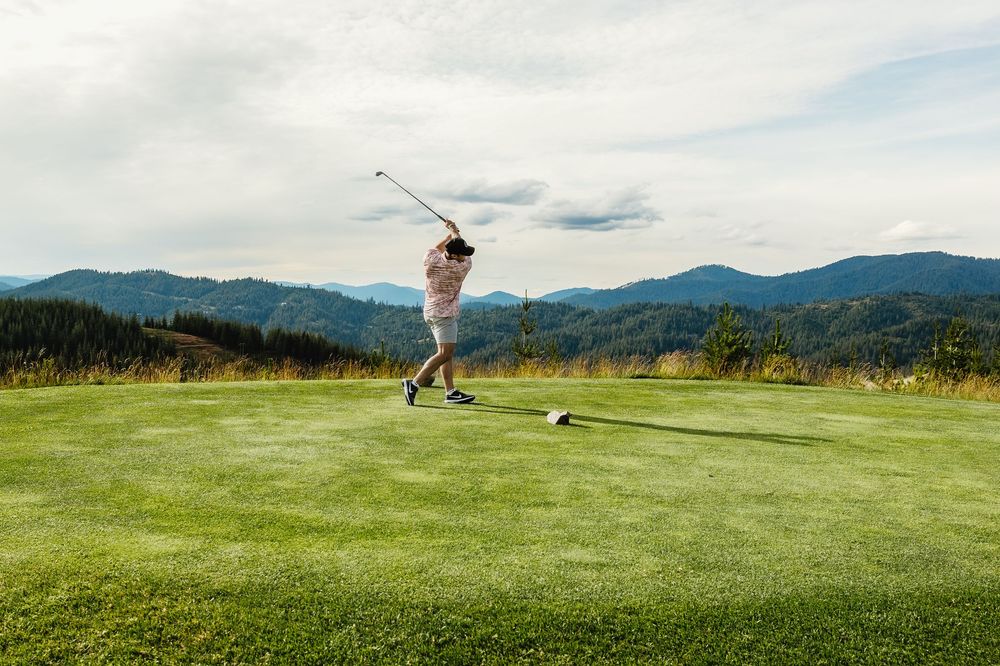 Golfer takes a swing on a scenic mountain course with lush green fairway and mountain range backdrop.