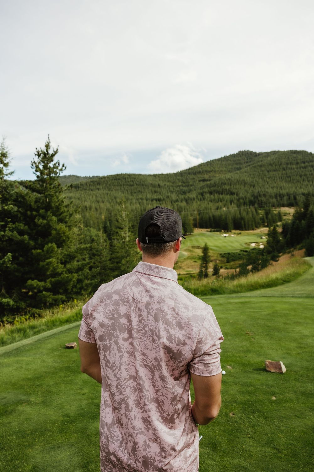 Golfer overlooking a scenic mountain golf hole surrounded by evergreen forests and hills.