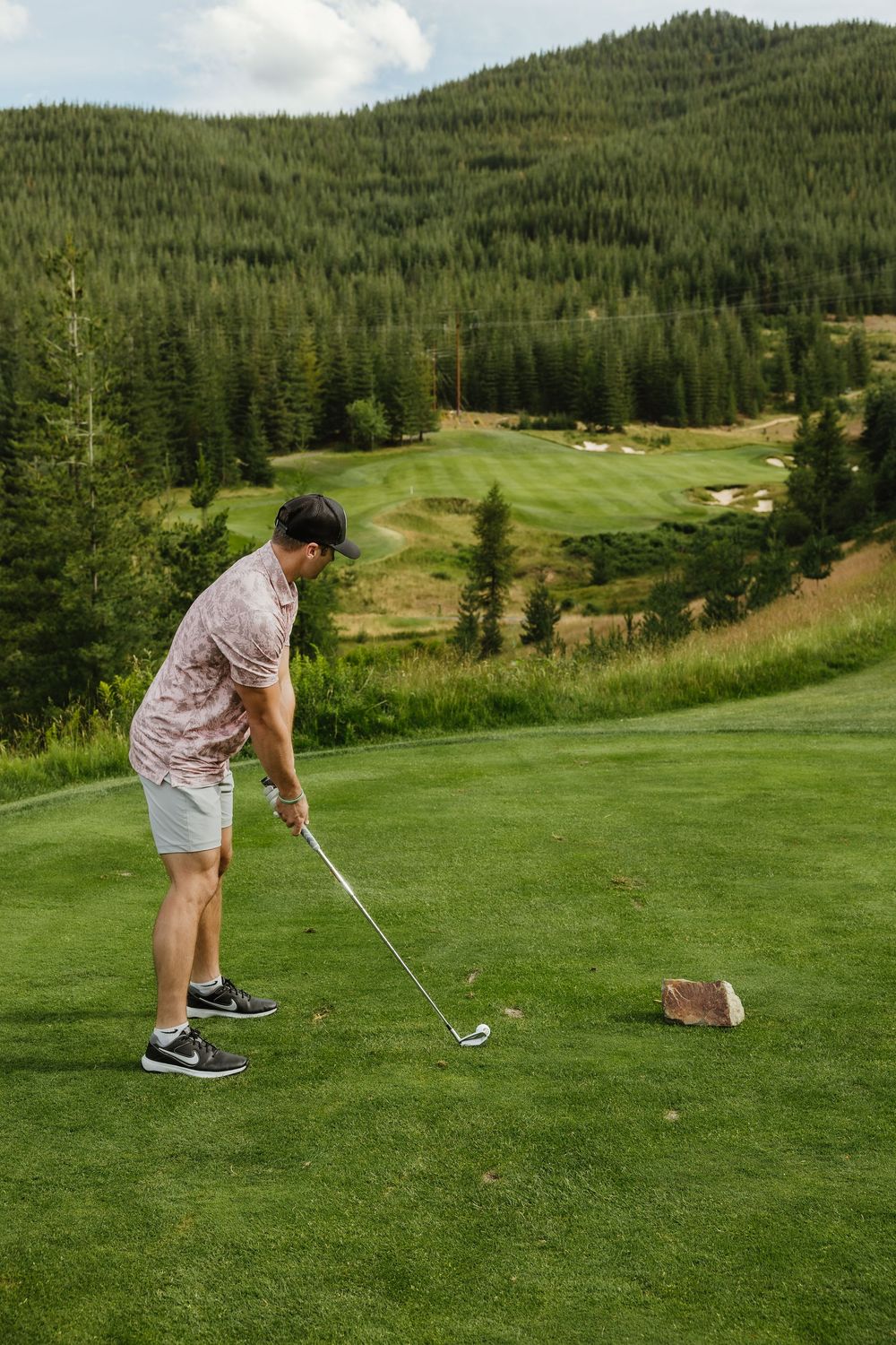 A golfer takes a swing on a scenic mountain course surrounded by evergreen forest slopes and rolling hills.