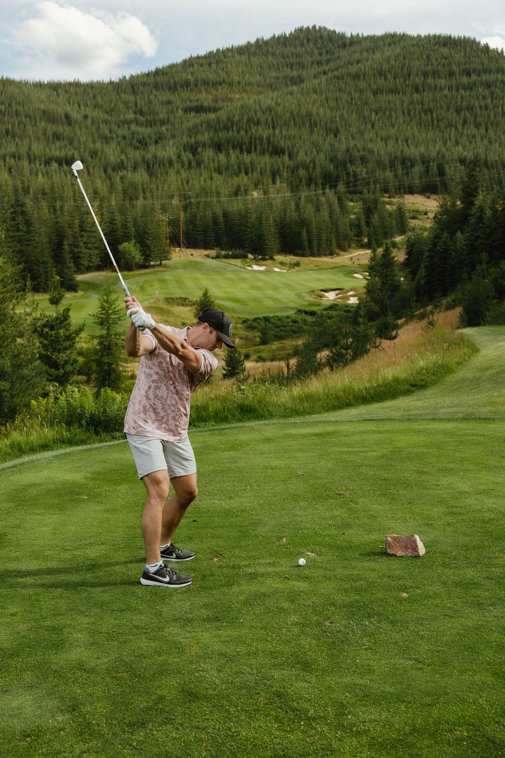 Golfer taking a swing on an elevated mountain course with scenic forest views and rolling fairways below.