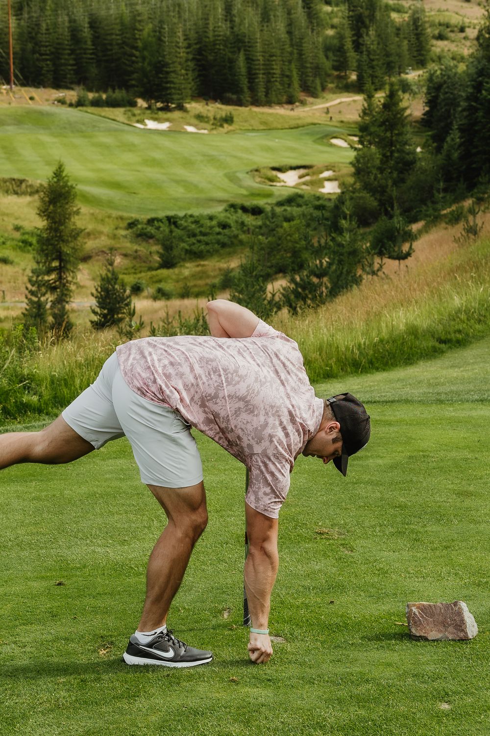 A golfer bends down to place a golf ball on the tee at a scenic mountain course with rolling hills and pine trees.