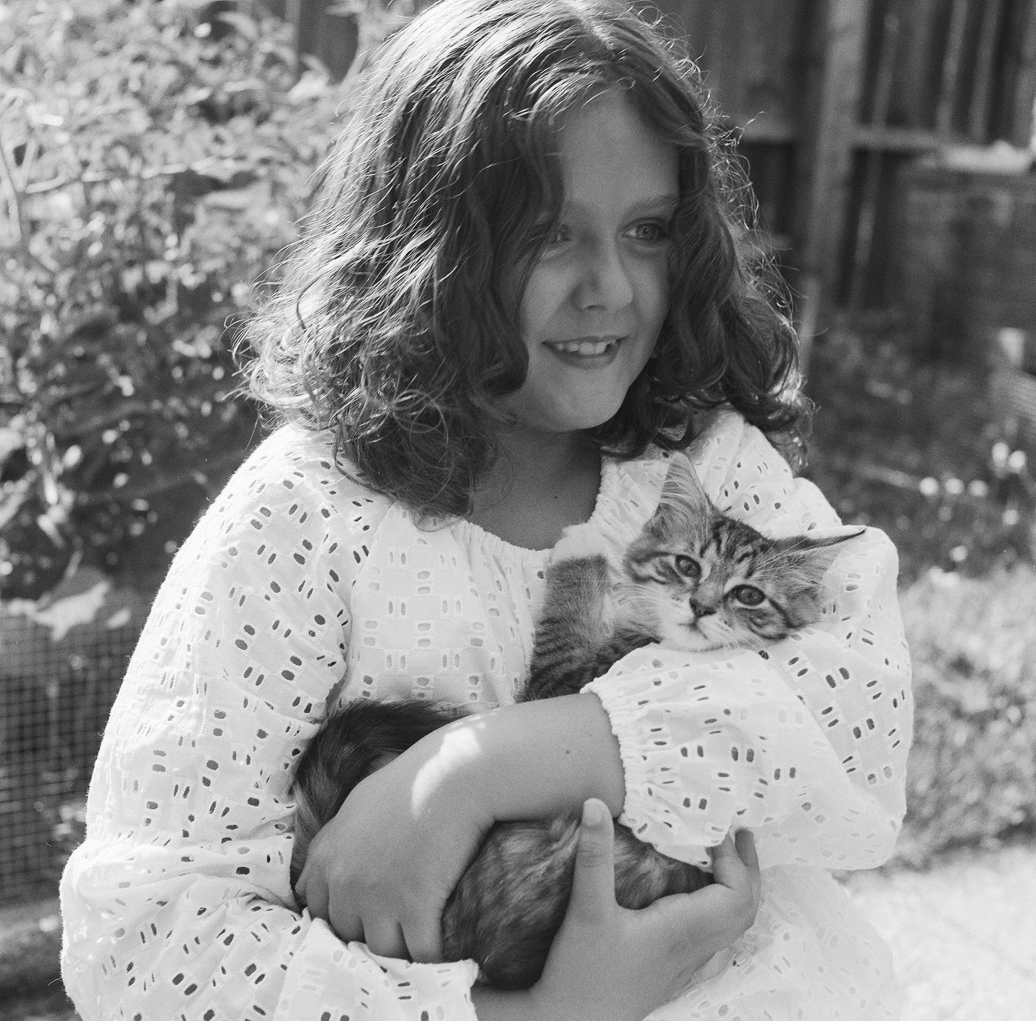 A series of black and white photographs showing someone in an eyelet dress holding and playing with a cat outdoors.
