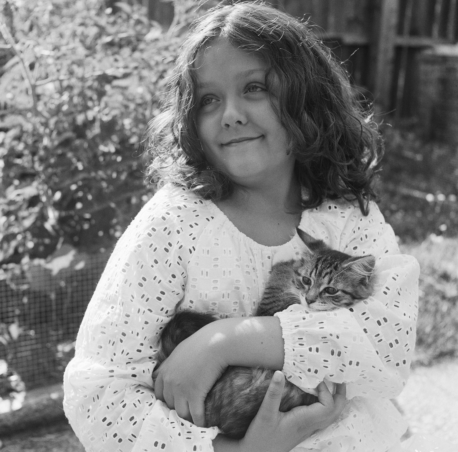A child in a white crochet sweater holds a contented gray tabby cat in this vintage black and white photograph.
