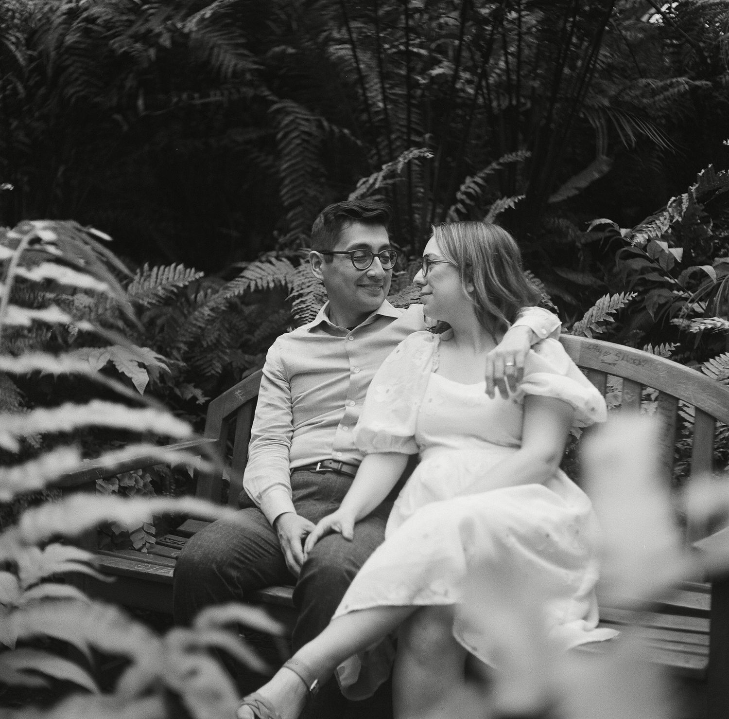 Romantic black and white wedding moment captured through garden foliage, showing couple sharing intimate embrace.