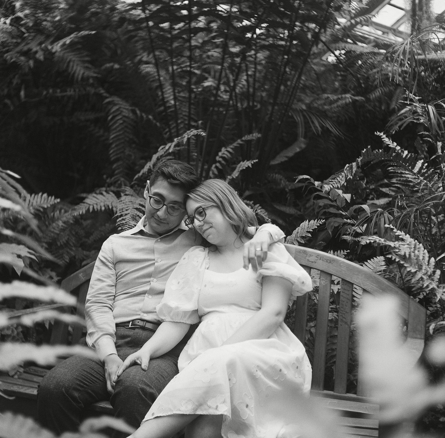 A romantic black and white photograph of a couple sitting close together on a bench surrounded by lush ferns.
