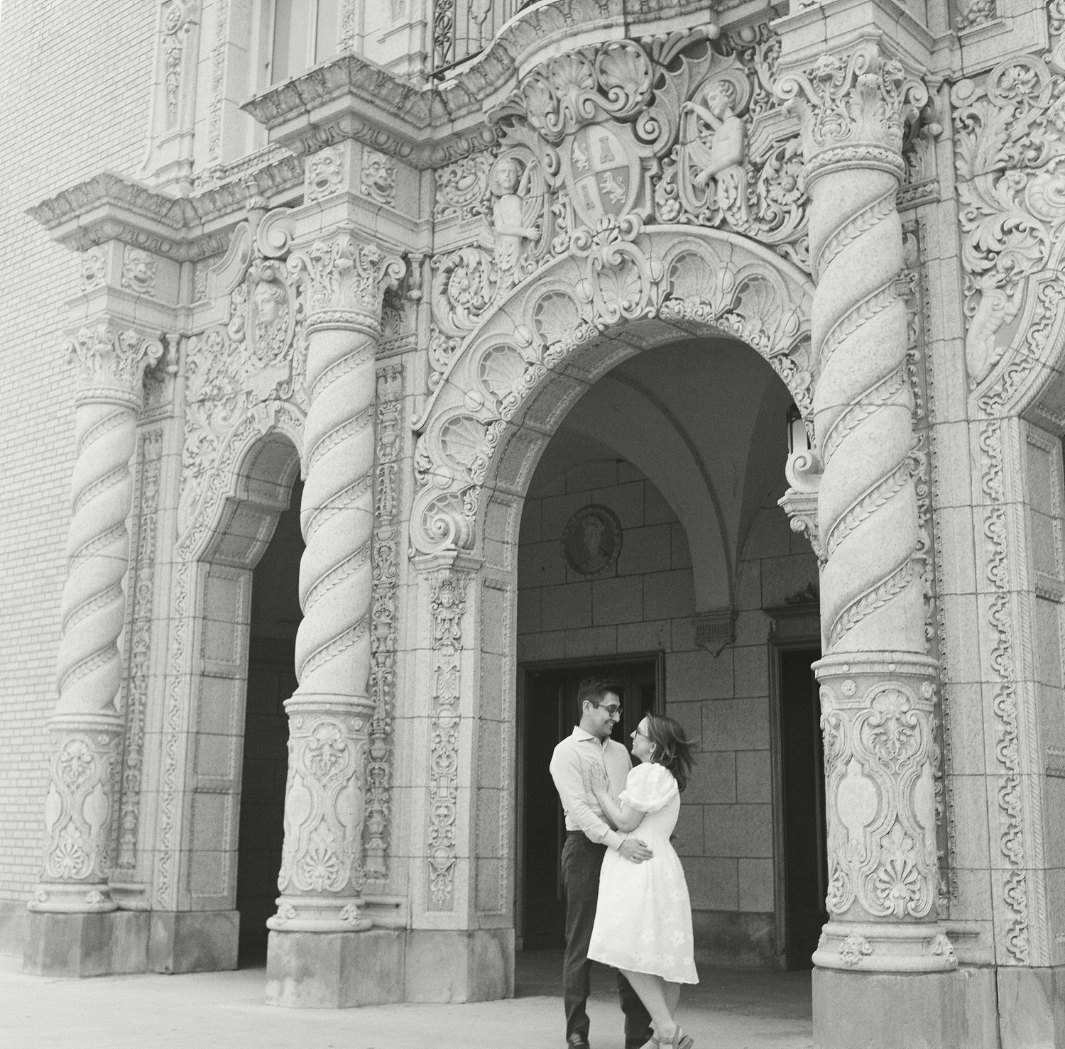 A couple embraces under ornate baroque archways with spiral columns and detailed stone carvings at a historic building.