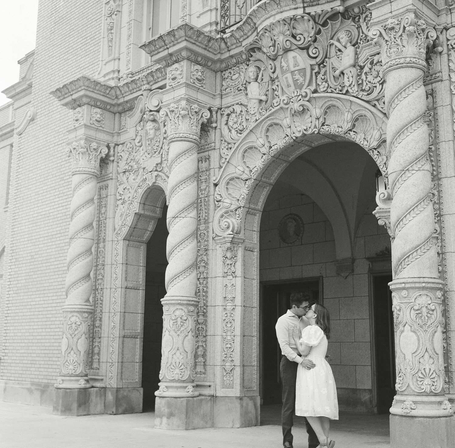 A couple poses for romantic wedding photos in front of an ornate white stone building with arched columns.