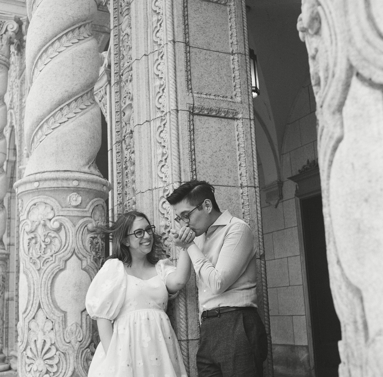 A black and white wedding photograph showing a couple and guests in a grand architectural setting with ornate columns.