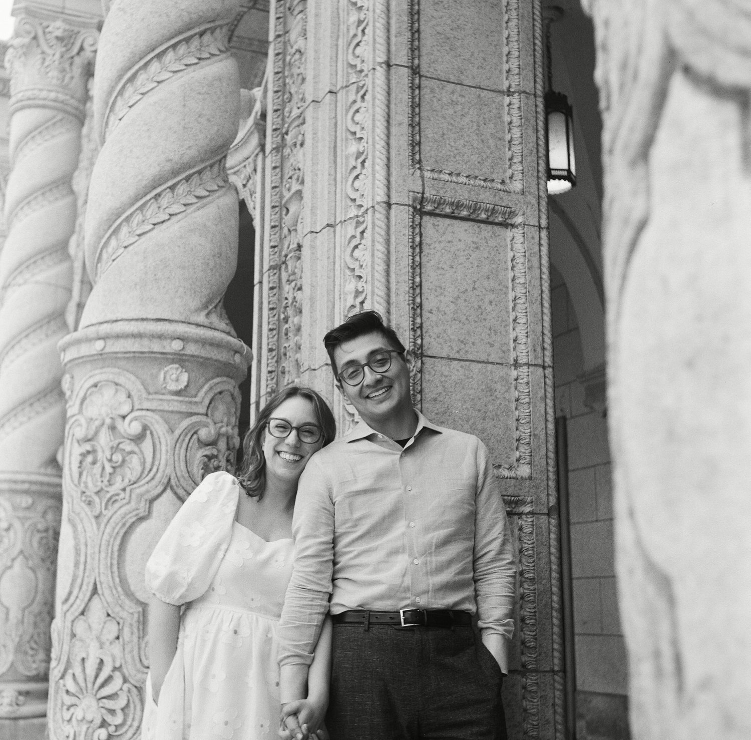 A couple stands between decorative stone columns with intricate carvings at a historic architectural site.