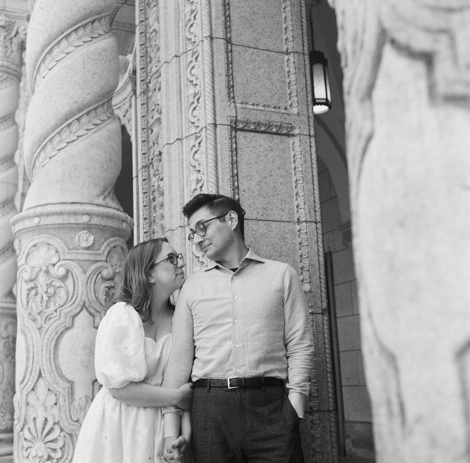 A black and white photo shows a couple embracing and leaning against an ornate stone pillar with carved details.