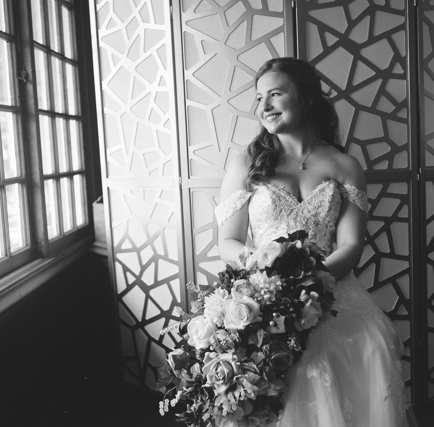 Black and white wedding photo of bride in elegant gown posing by window with natural light and floral bouquet.
