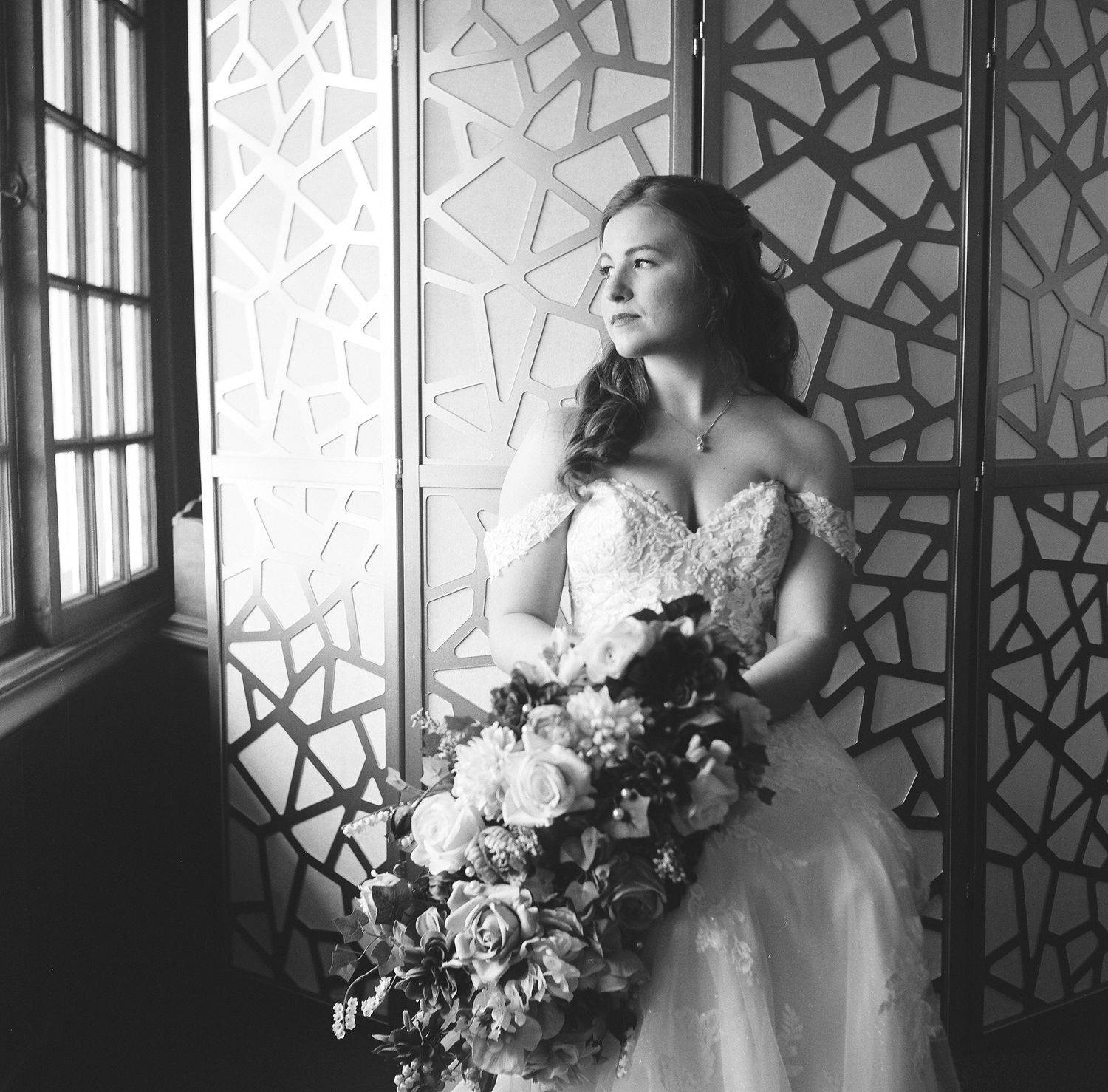 A bride in a white gown poses near a decorative window screen while holding a luxurious floral bouquet.