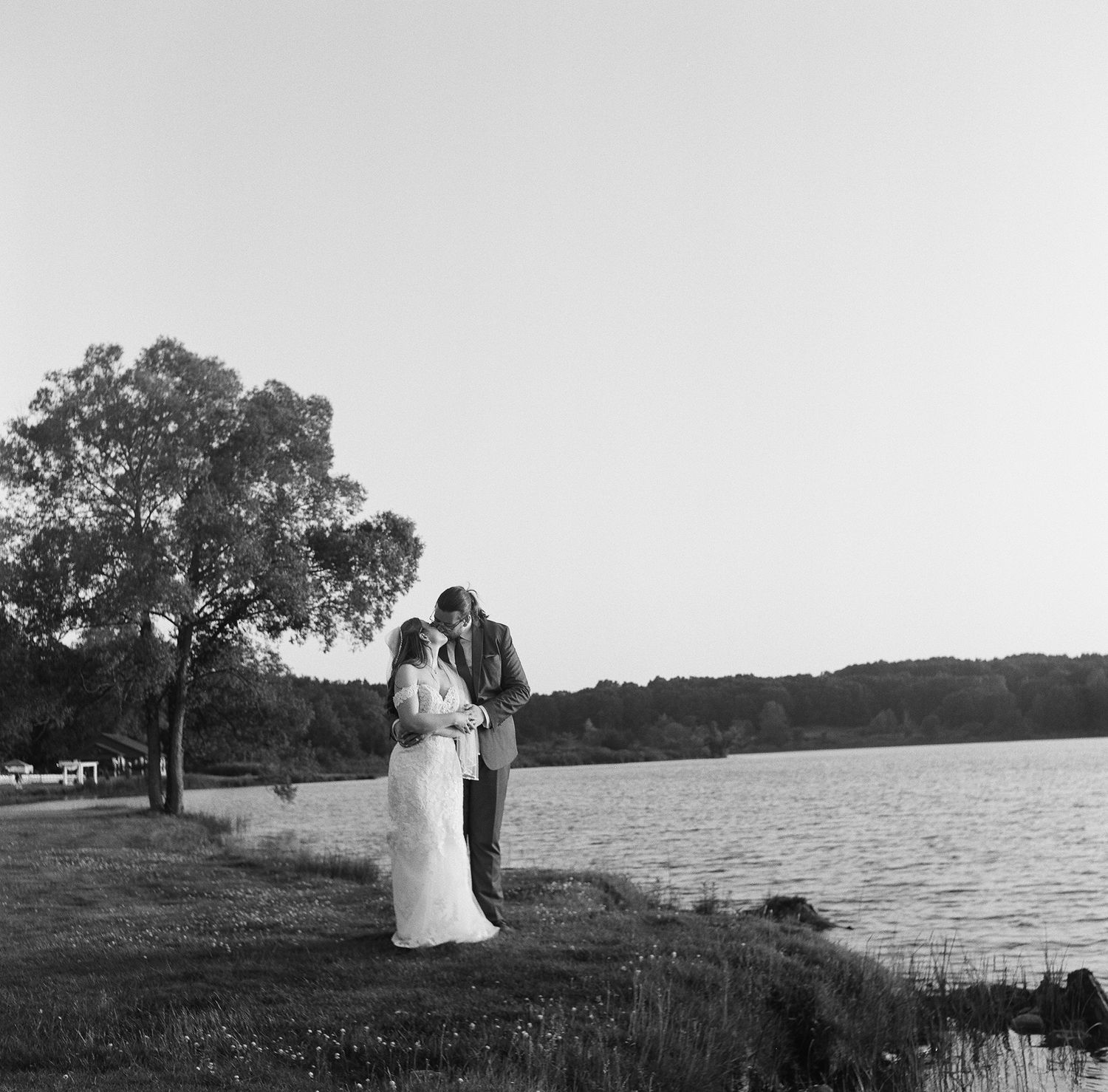 A couple shares a romantic moment by a serene lake at sunset with trees silhouetted in the background.