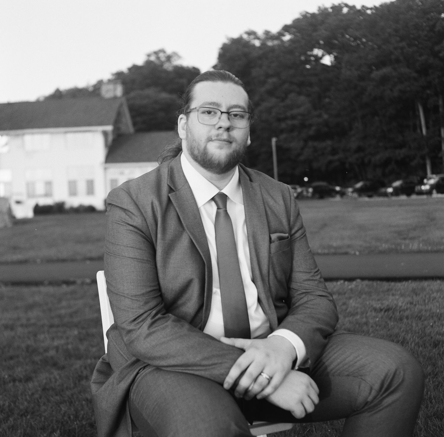 A groom in a dark suit relaxes on grassy grounds during an outdoor wedding photoshoot.