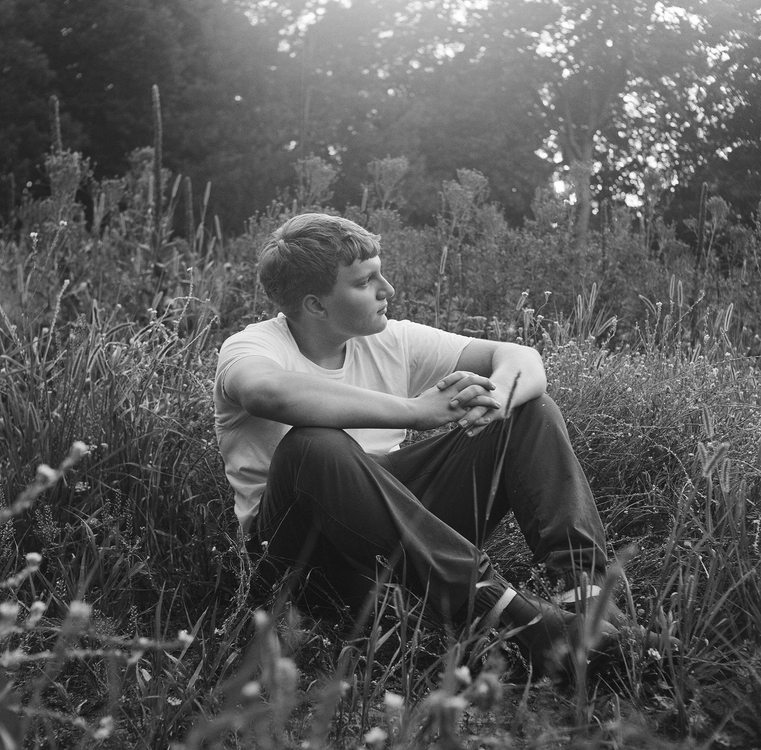 Black and white photograph of a person sitting thoughtfully in a meadow of tall grass on a sunny day.