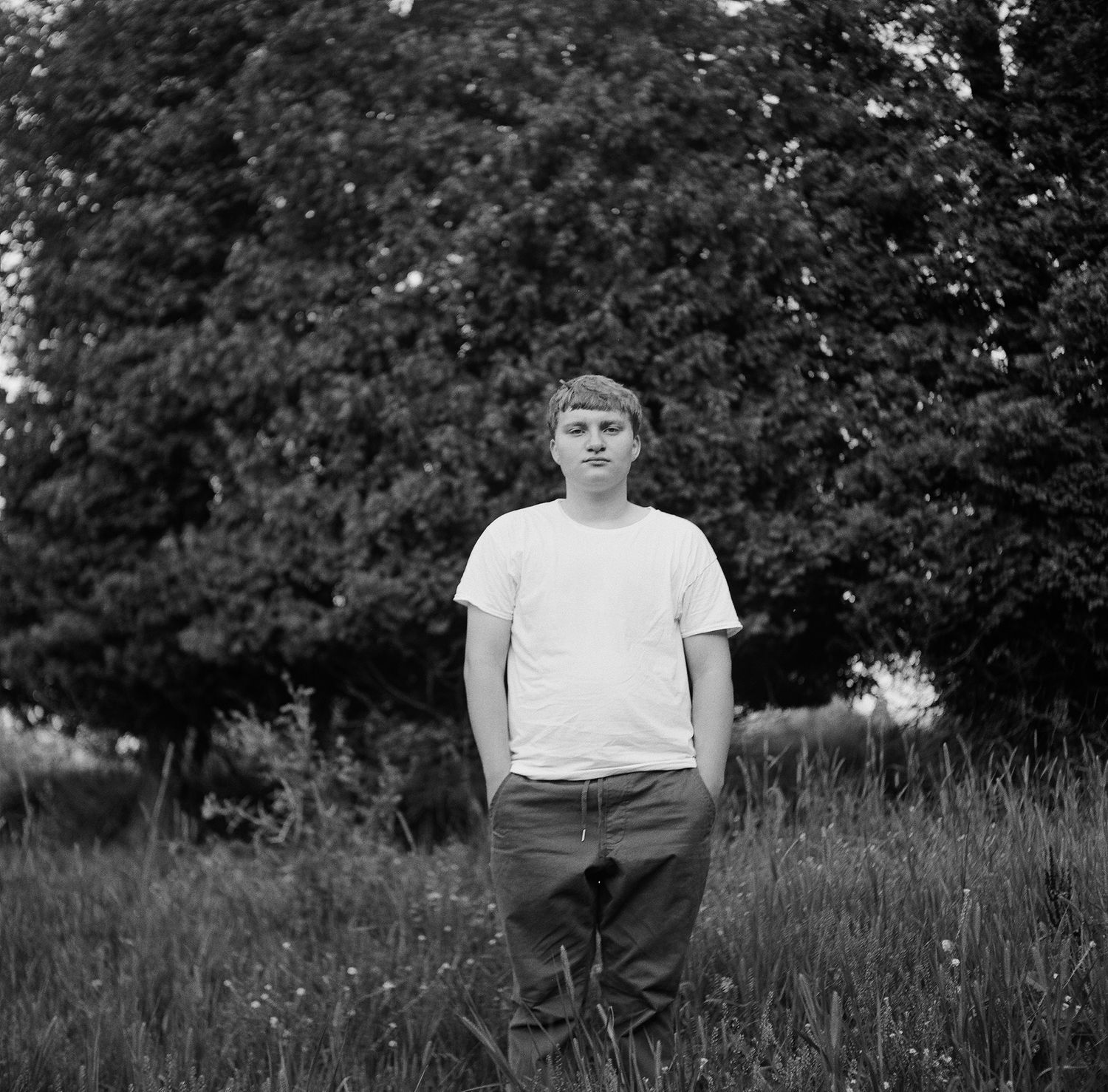Black and white portrait of someone in a white t-shirt standing outdoors in front of leafy trees and foliage.
