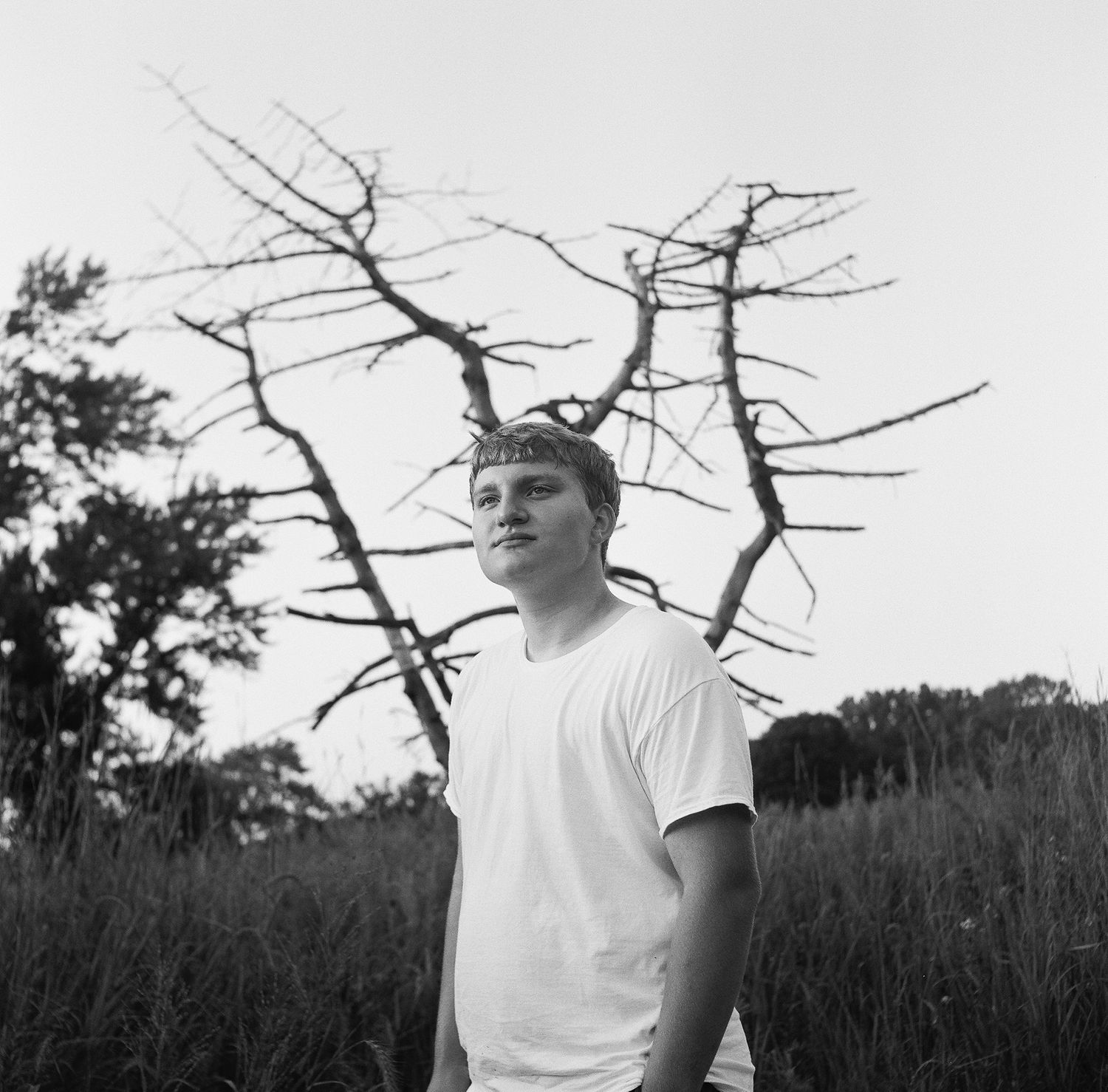 Black and white portrait of someone wearing a white t-shirt in a natural outdoor setting with bare trees.