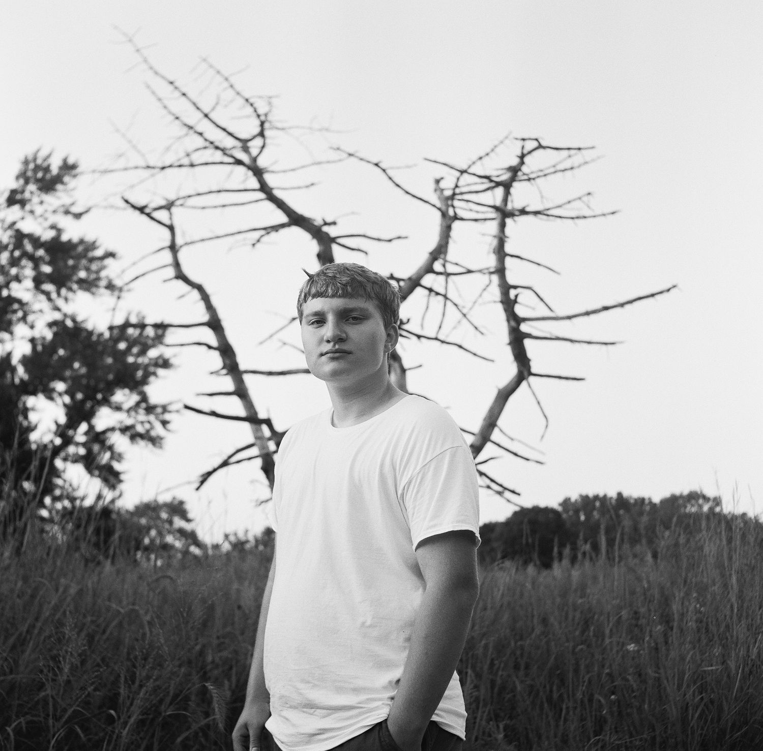 Black and white portrait of someone wearing a white t-shirt in a natural outdoor setting with bare trees.