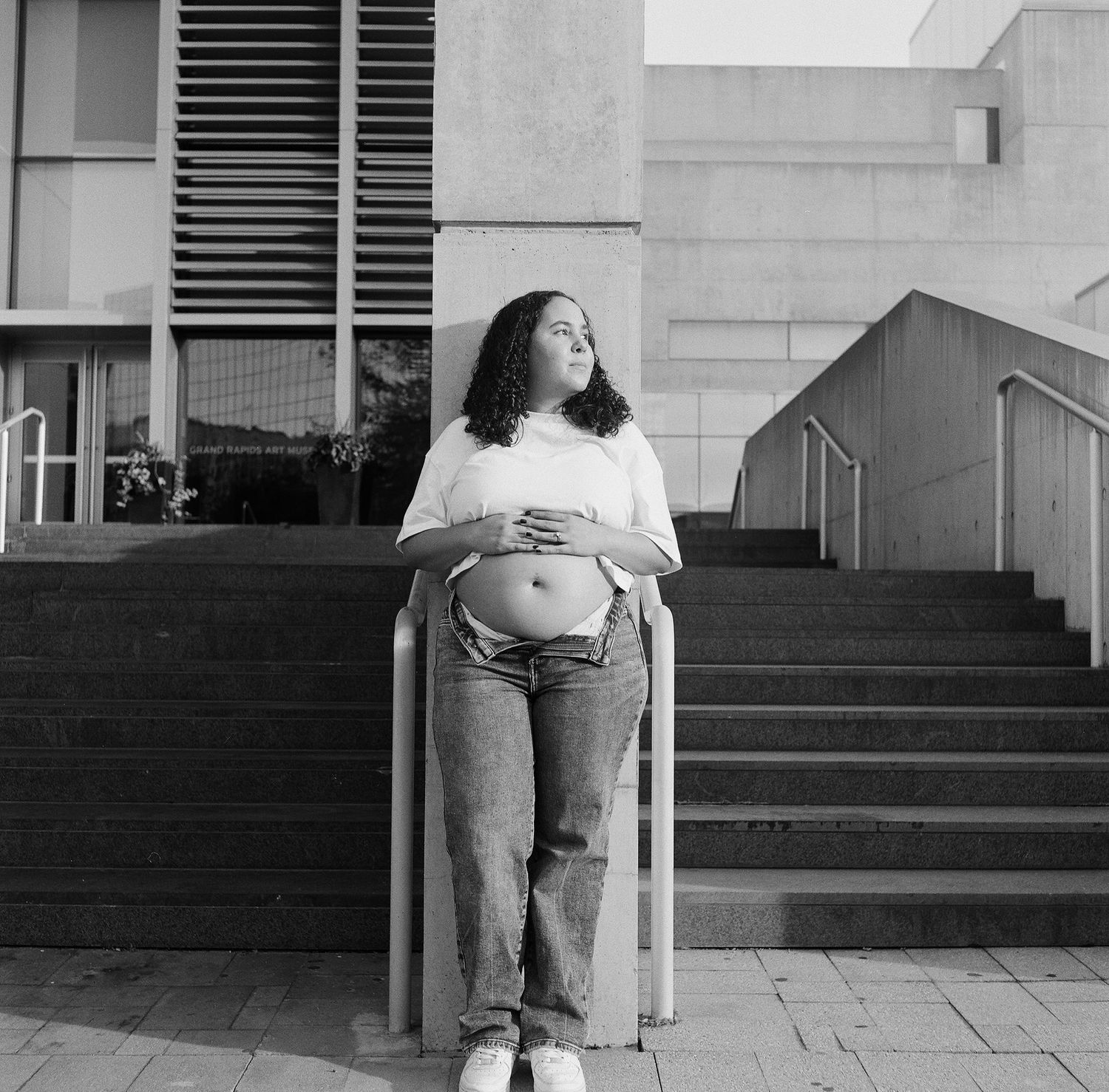 Black and white photo of a pregnant woman in casual wear sitting on a concrete staircase outdoors.