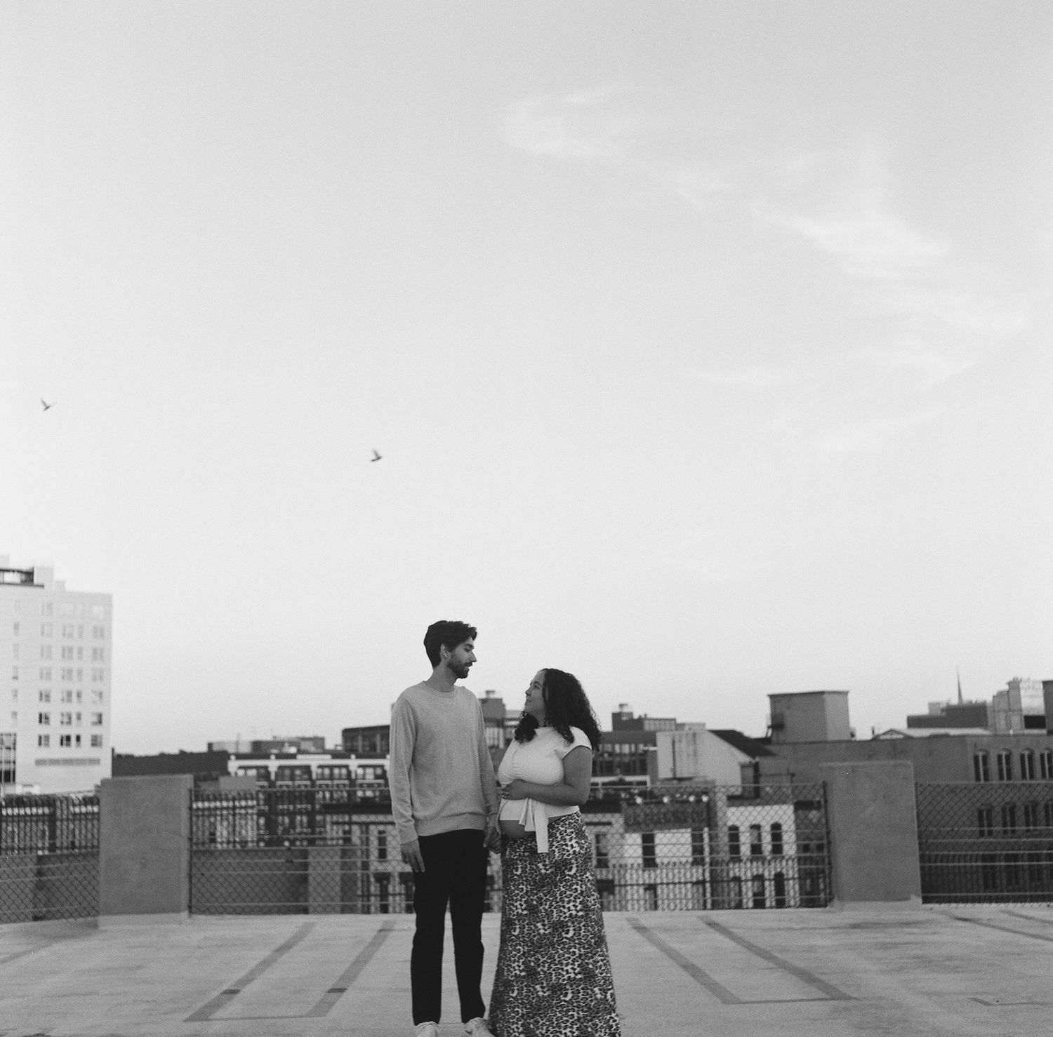 Two people stand on a rooftop overlooking an urban skyline of buildings and water towers at dusk.