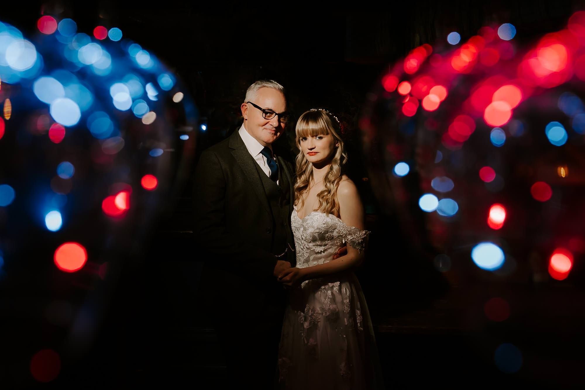 A couple dances together in a dark room with red and blue bokeh lights creating a romantic atmosphere in the background.
