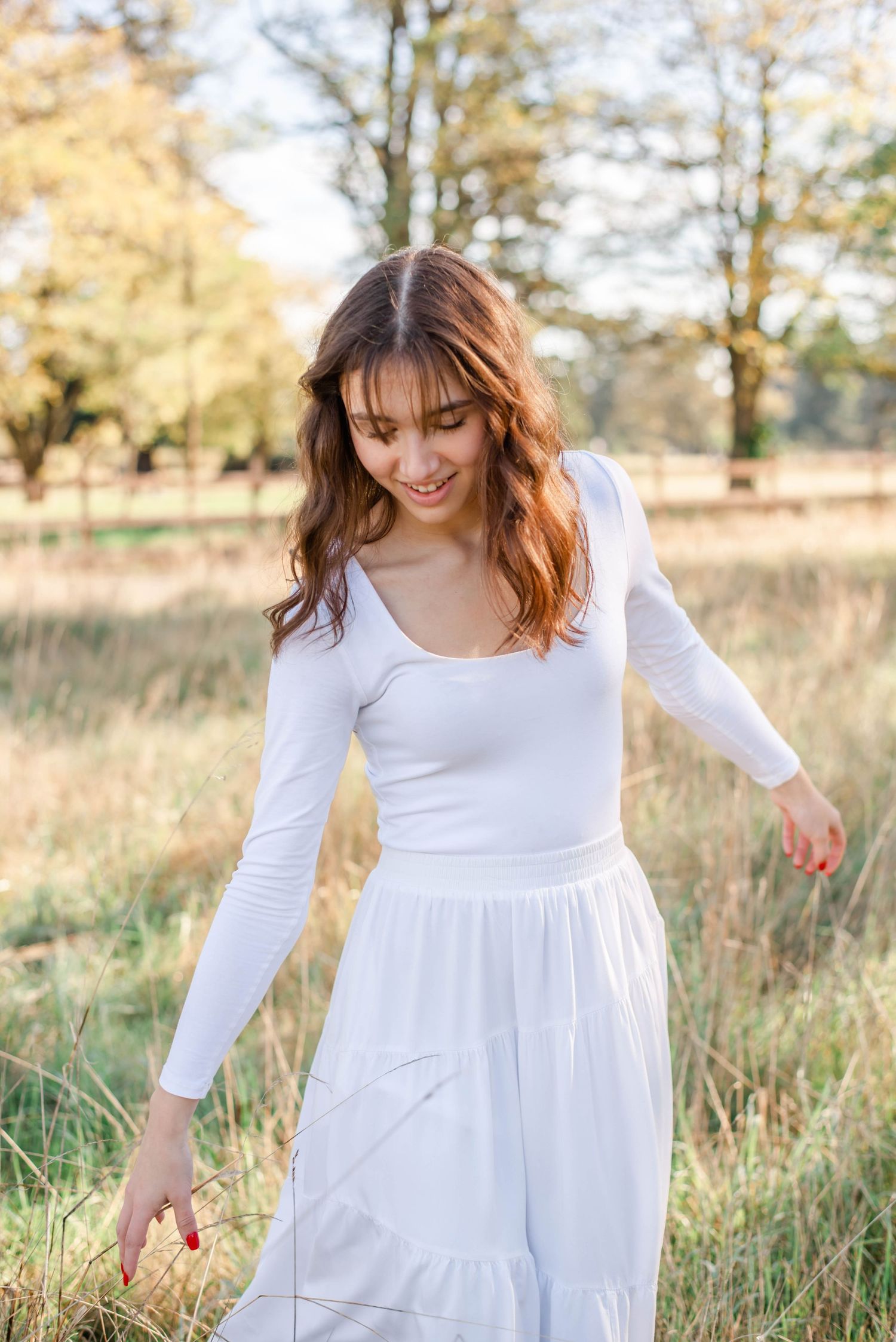 Woman in flowing white dress walks through golden field at sunset during outdoor portrait session.