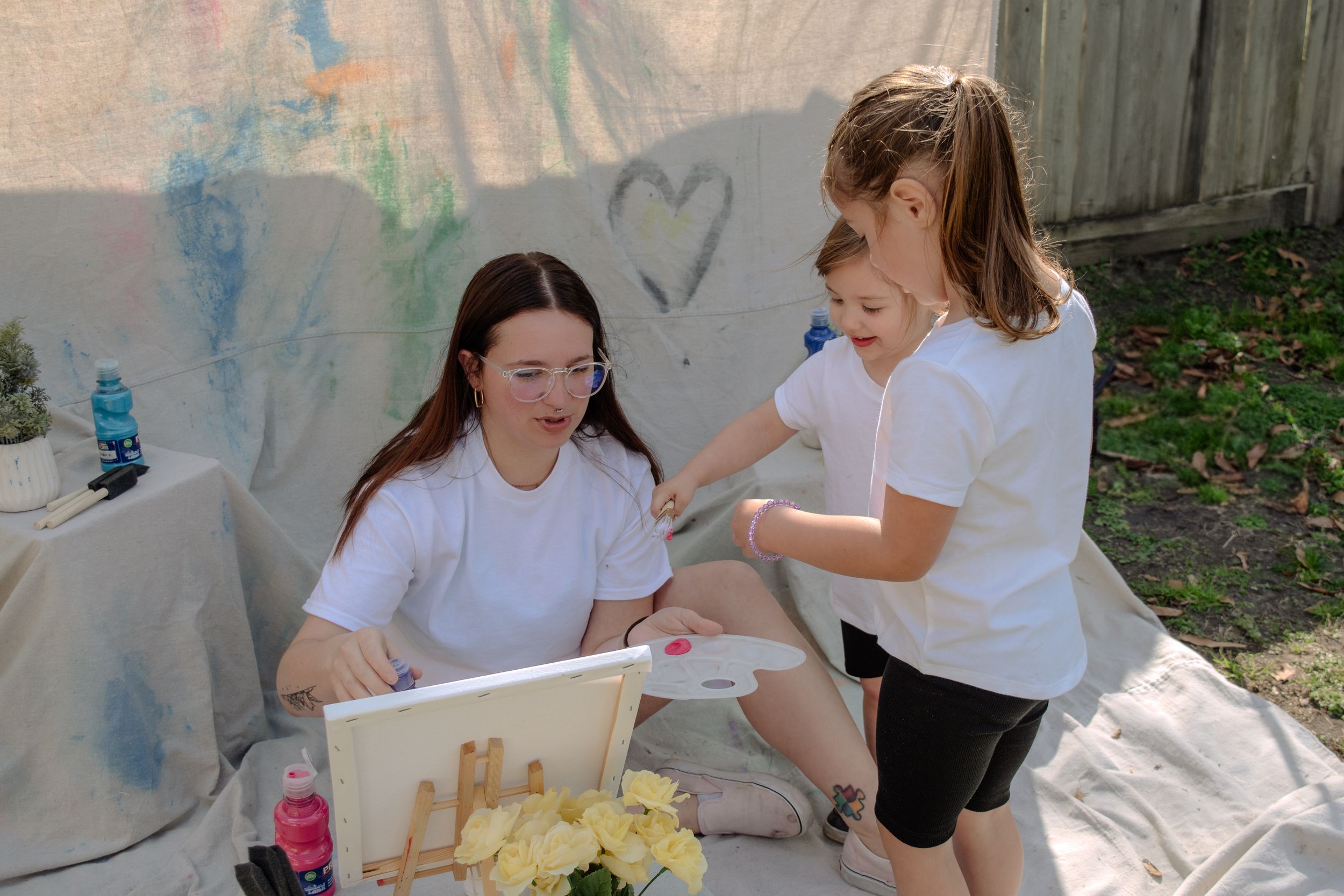 Two students in white shirts work together on an art project outdoors near a fence.