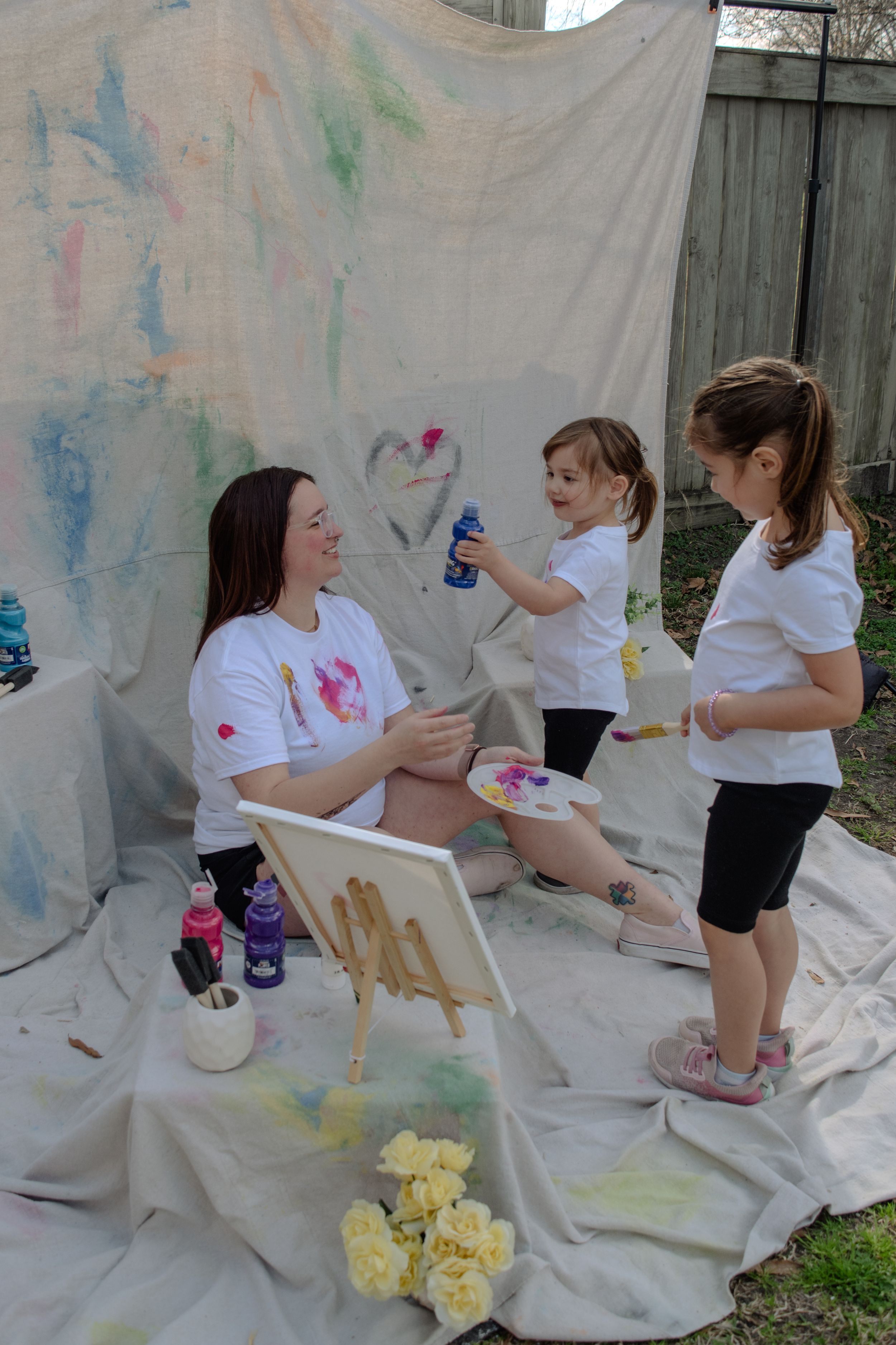 Children in white shirts create art on a white backdrop while sitting on the ground with paint supplies.