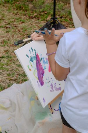 Children display their colorful handprint artwork on white paper while sitting outside on a sunny day.