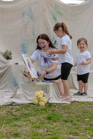 Children in white shirts create colorful art on a large canvas spread outdoors on grass.