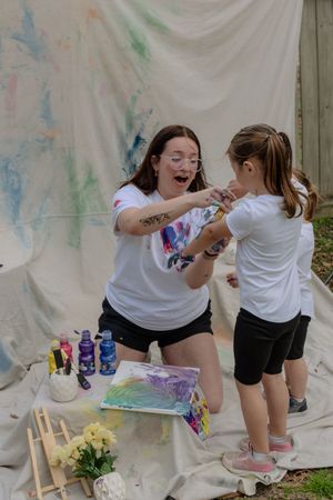 Two people in white shirts create colorful tie-dye art together outdoors against a white backdrop with paint supplies.